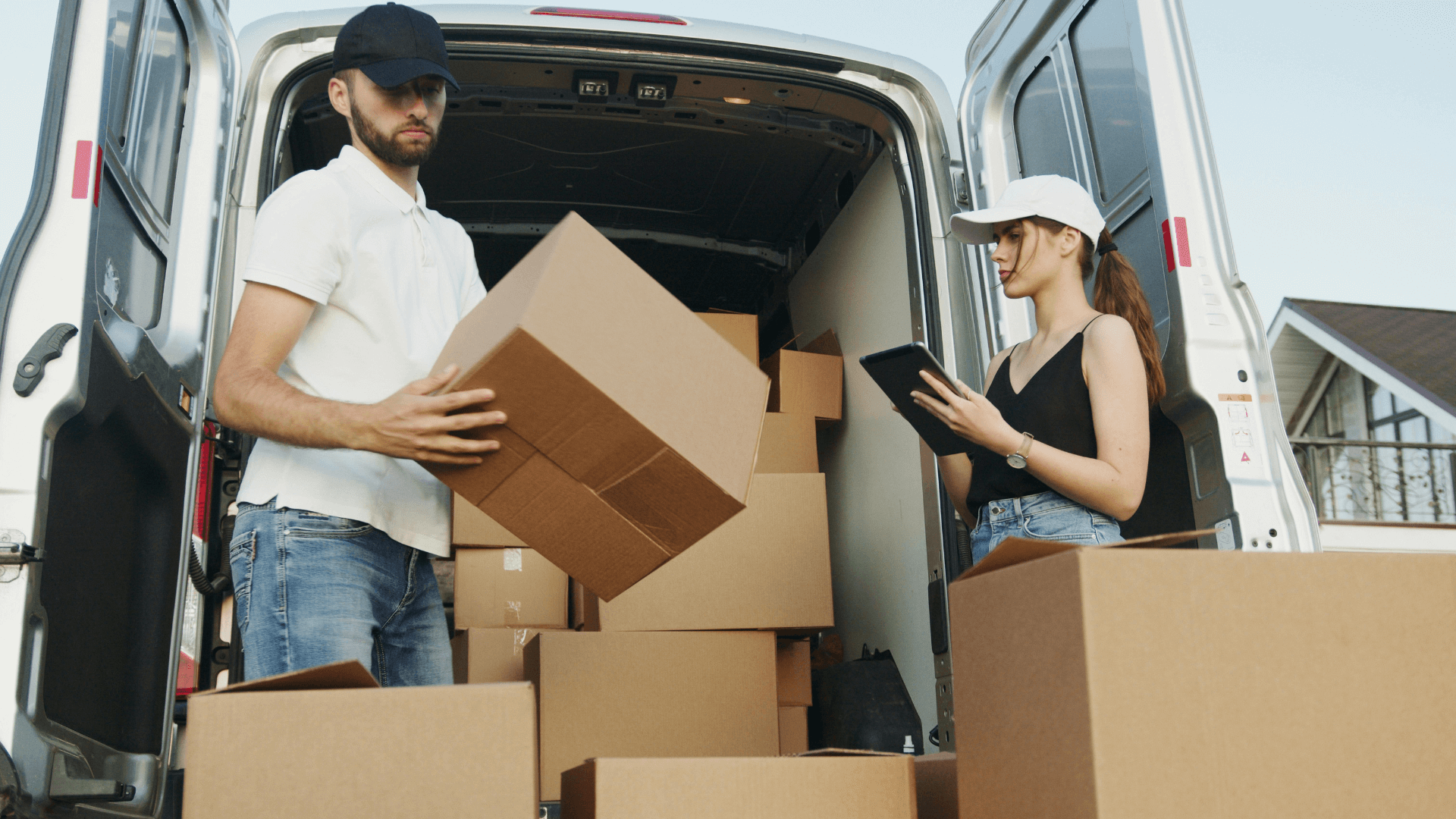 Two workers loading cardboard boxes into a delivery van while checking a tablet