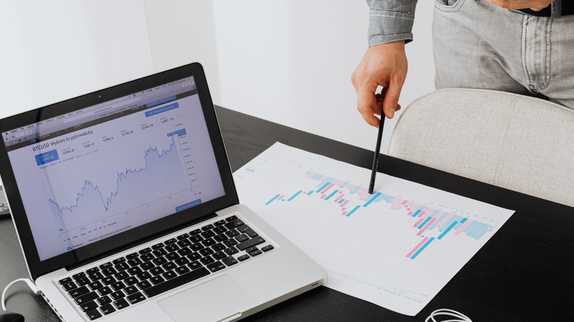 Hand holding a pen over a printed sales chart beside a laptop showing graphs