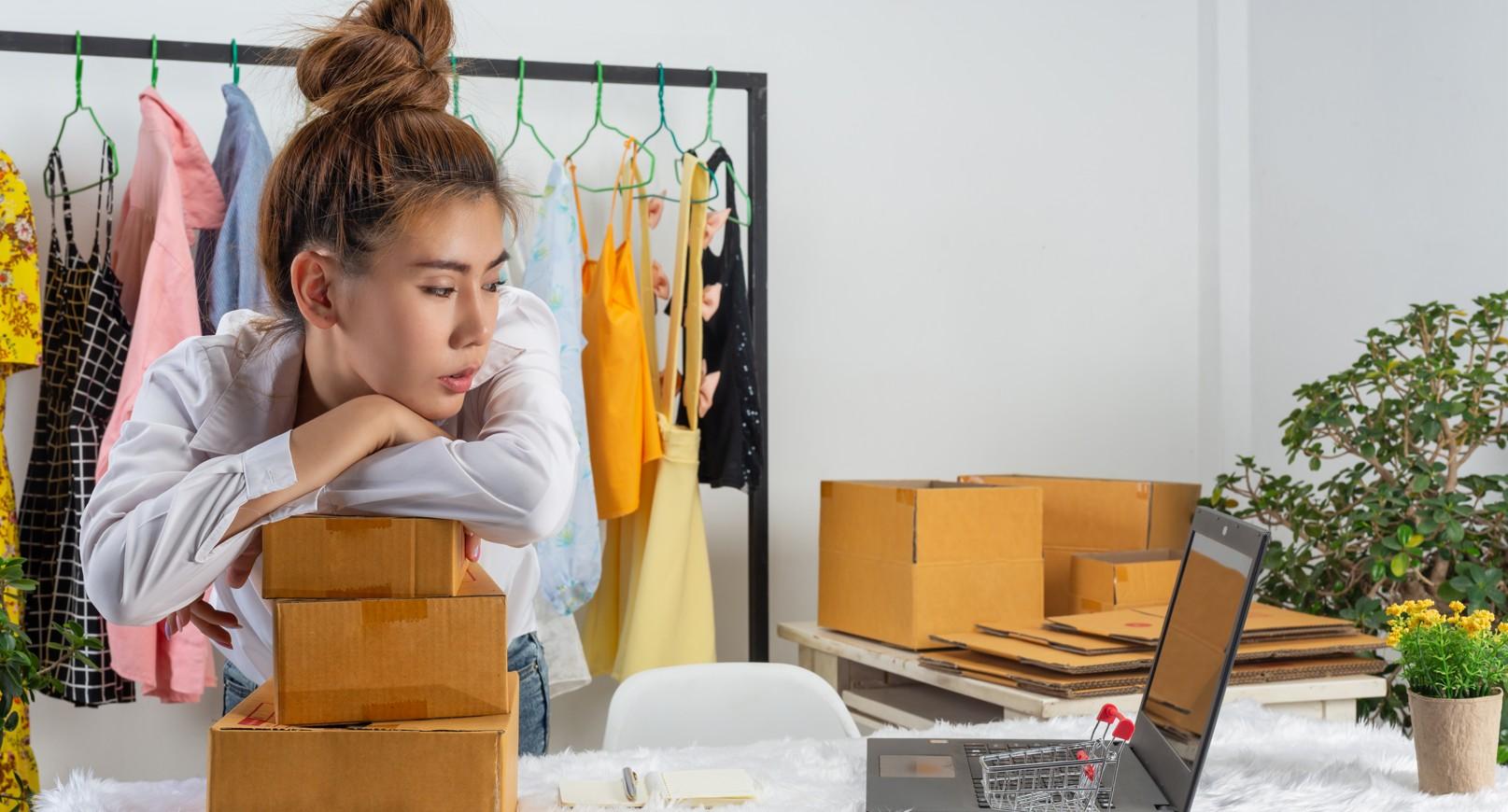 Seller at a laptop surrounded by parcels and a clothing rail, looking discouraged