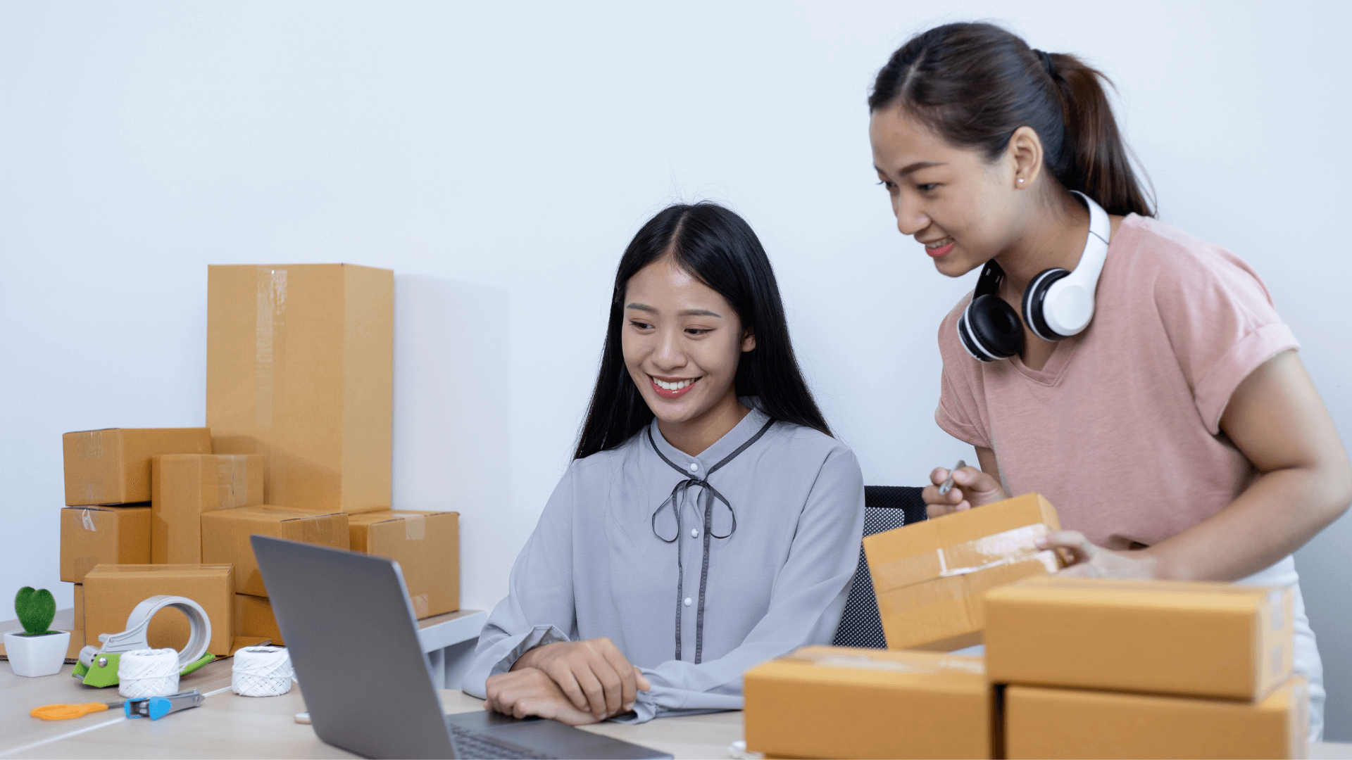 Two sellers packing parcels at a laptop surrounded by boxes